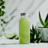 A bottle of aloe juice, greenish tint, displayed on a white surface, with aloe vera plants and sliced aloe in the background.