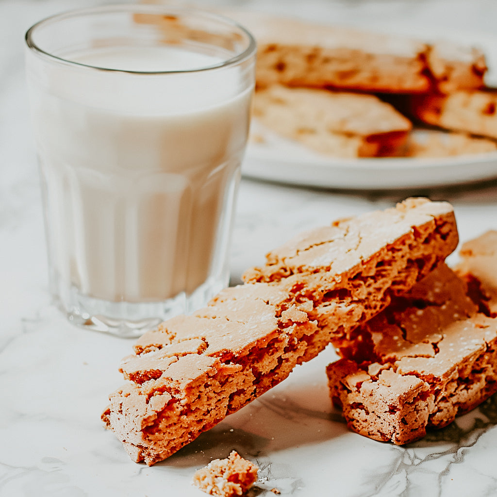 A glass of milk accompanied by several biscotti cookies on a white surface.