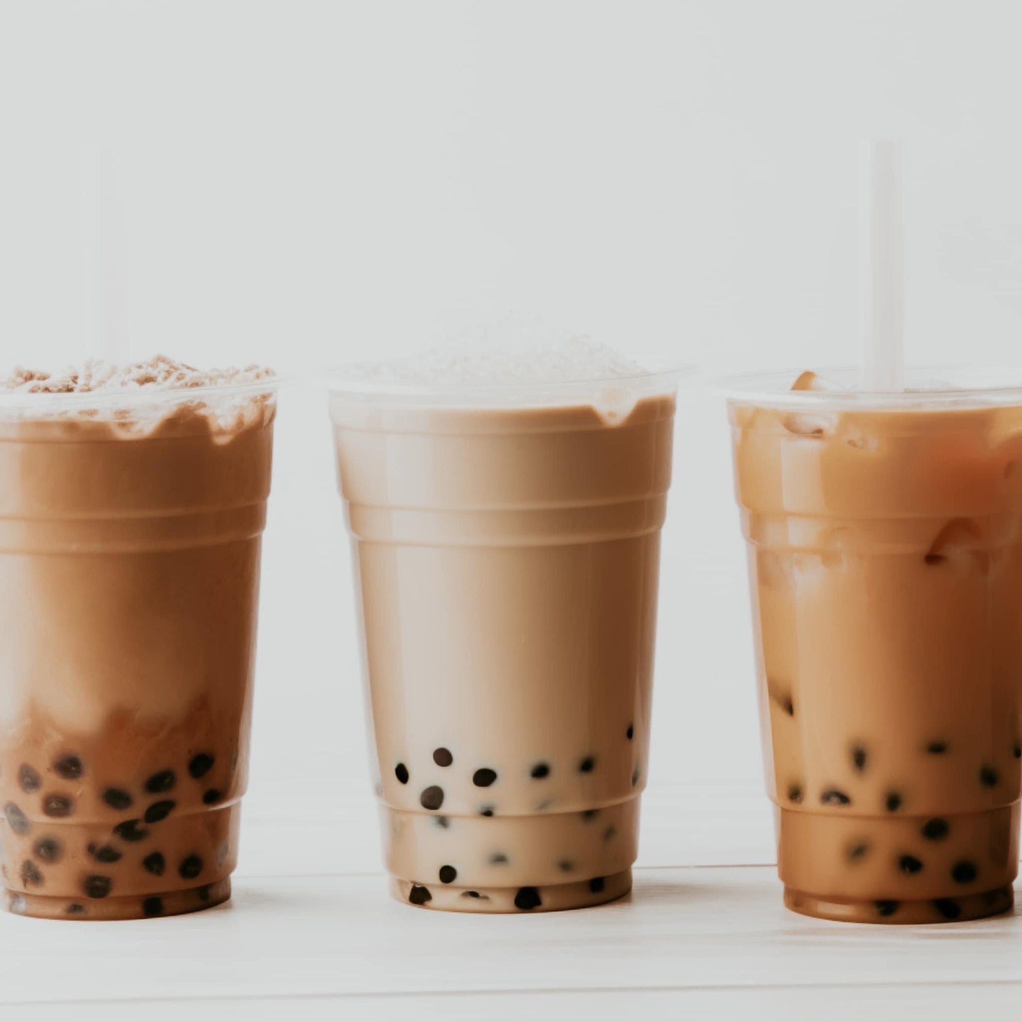 Three cups of bubble tea in plastic take-out cups, arranged side by side on a white surface. Each cup contains visible boba pearls and varies in color, suggesting different flavors.