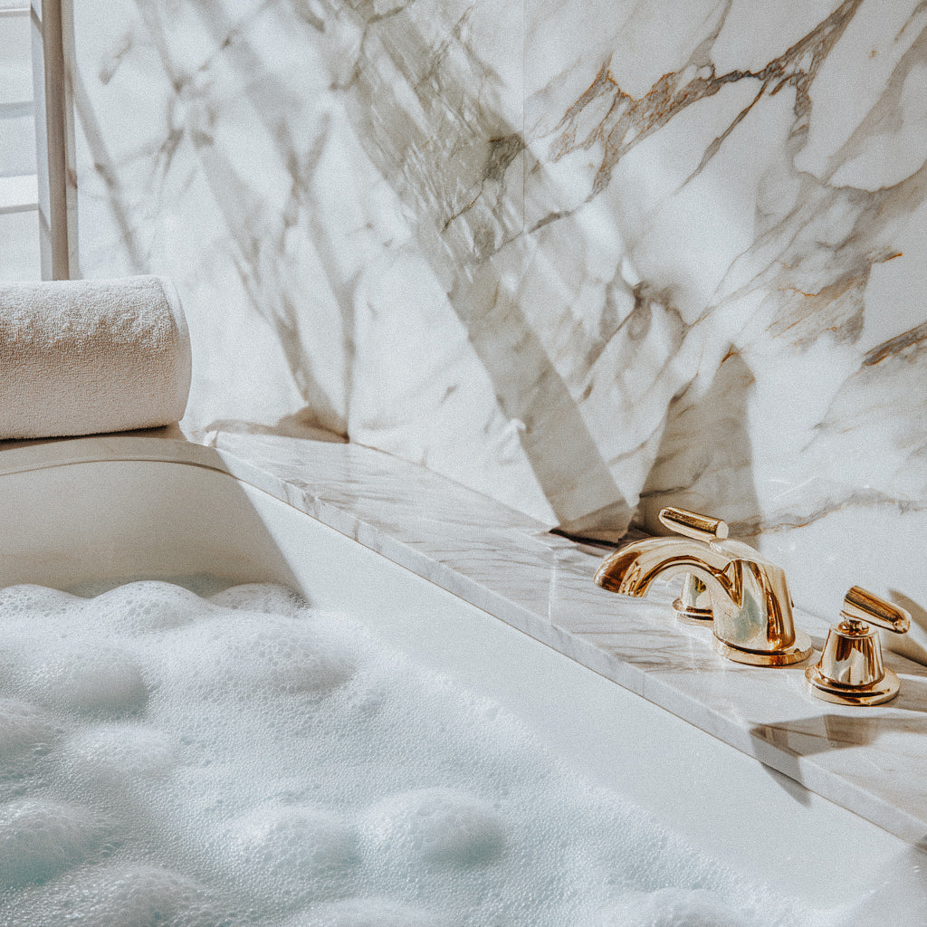 A bubble bath running in a bathtub with white marble walls, featuring gold-colored faucets and a stack of white towels at the edge of the tub.