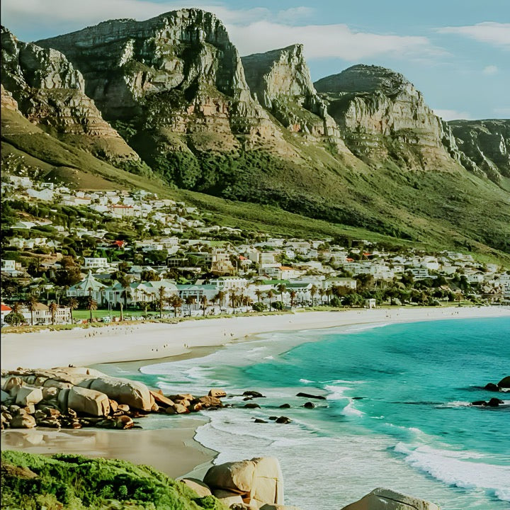 A scenic view of Cape Town featuring a beach with clear blue water, rocky outcrops, and a cityscape with green mountains in the background.