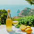 A bottle and a glass of limoncello placed on a table with a view of the ocean and lemon fruits in the foreground.