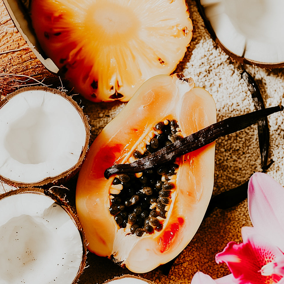 A photo of a halved papaya, a coconut cut open, and vanilla bean, arranged with a flower in the background.