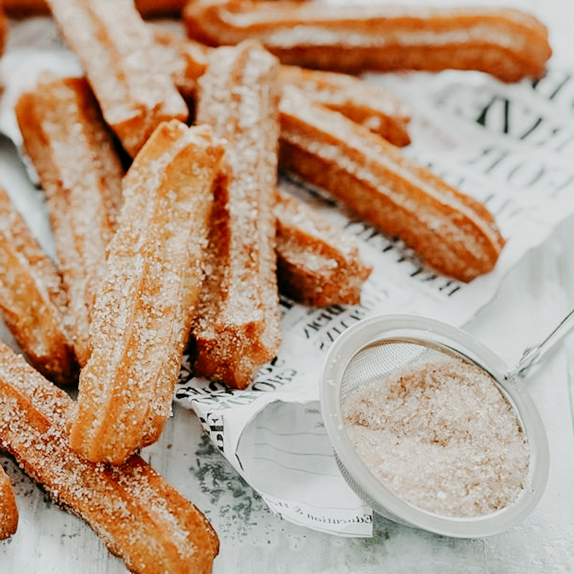 A batch of sugar-coated churros, freshly made and sprinkled with cinnamon, displayed on a paper-lined plate with a small bowl of sugar on the side.