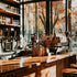 Interior view of a coffee house featuring a counter with coffee machines, glasses of coffee, and decorative items, with a window in the background showing trees and sky.