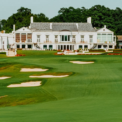 A well-manicured golf course with a country club house in the background, featuring large windows and a white exterior.