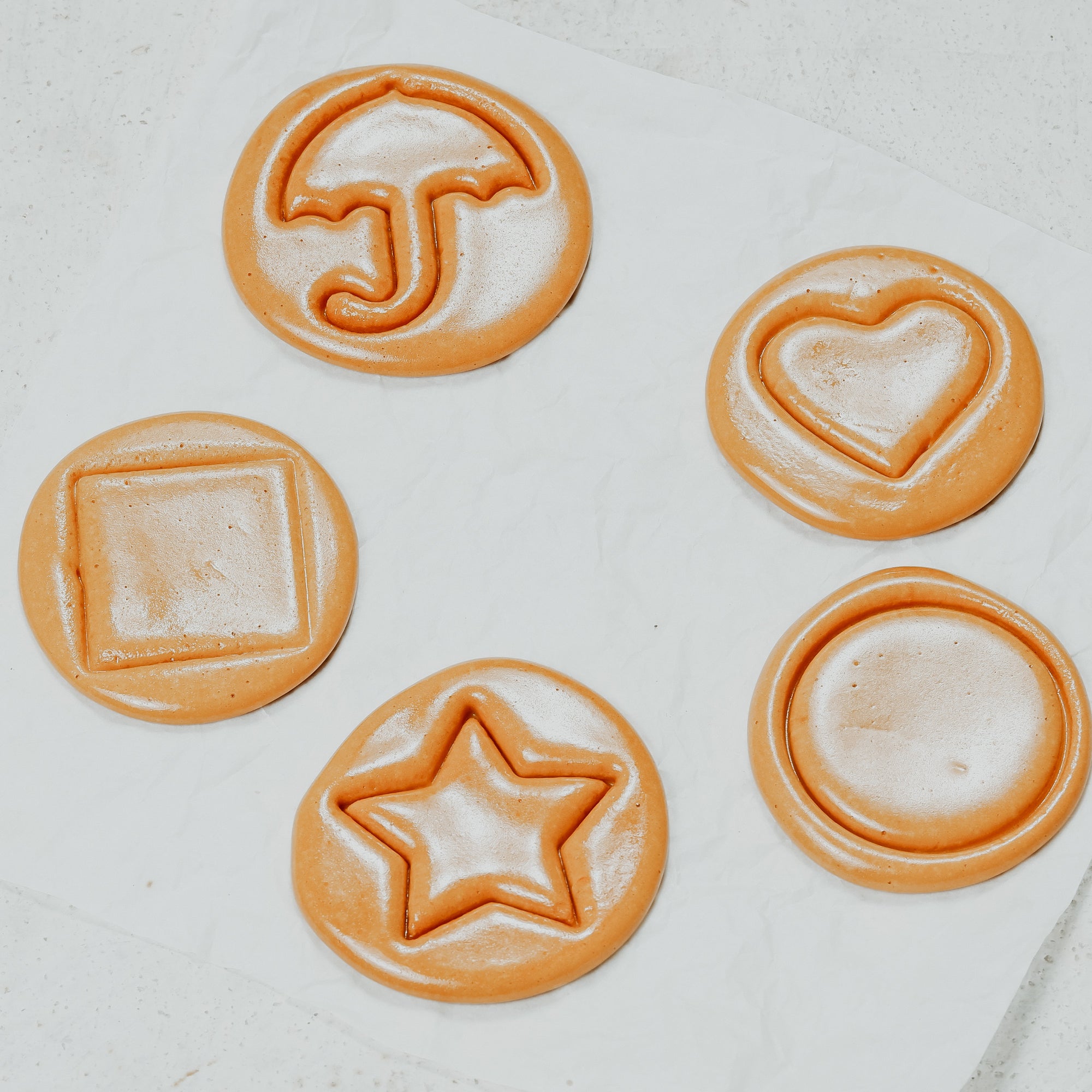 Four circular pieces of Dalgona Candy with a caramelized sugar scent, each with a different playful shape: an umbrella, a heart, a square, and a star, displayed on a light background.
