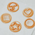 Four circular pieces of Dalgona Candy with a caramelized sugar scent, each with a different playful shape: an umbrella, a heart, a square, and a star, displayed on a light background.