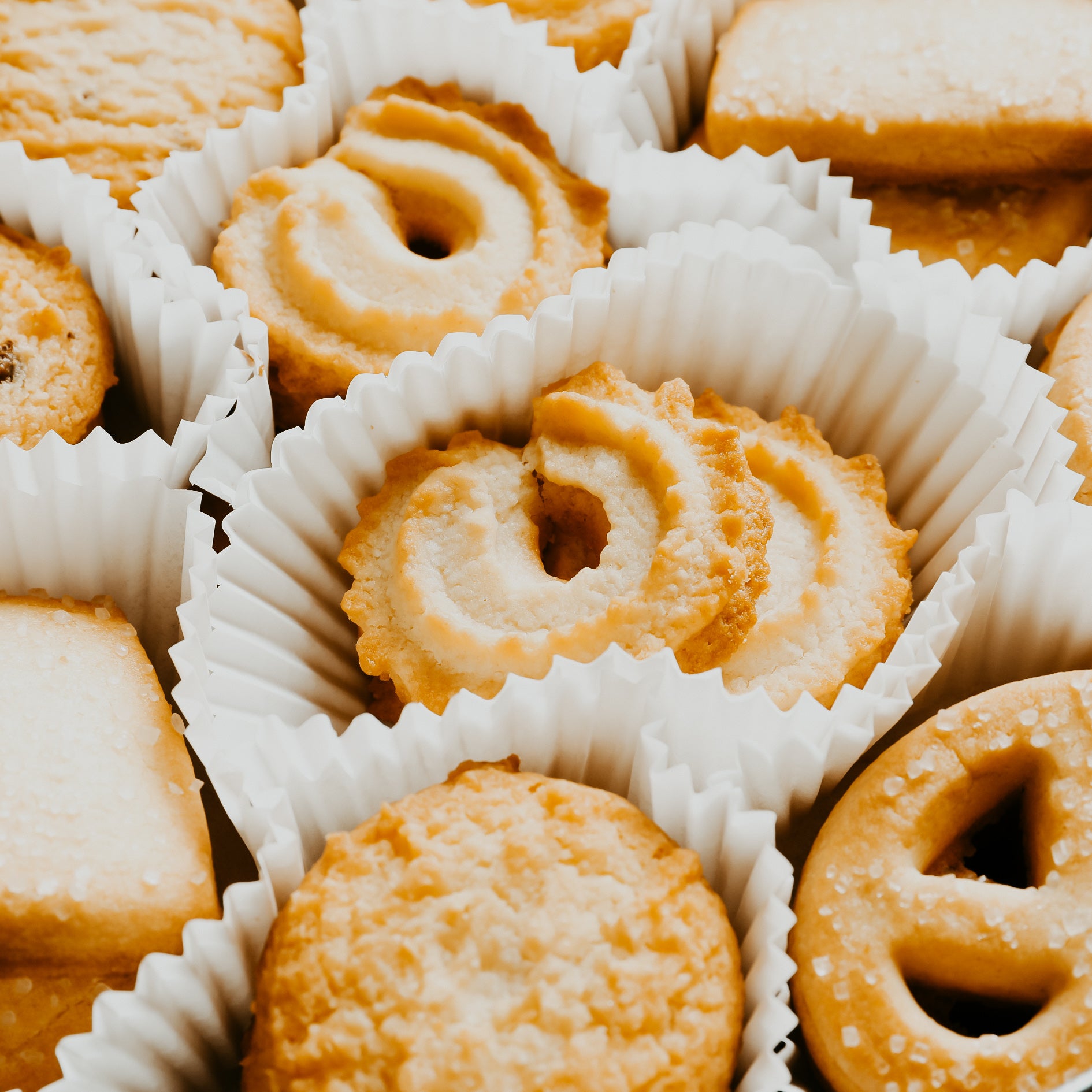 A close-up of Danish butter cookies arranged in white paper liners, with a creamy beige color and a slightly golden brown tint.