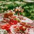 A picnic setup with a variety of food including fruits, bread, and a floral arrangement, on a checkered red and white blanket.