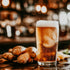 A glass of ginger beer with a foamy head, alongside fresh ginger roots, with a blurred background suggesting a bar or kitchen setting.