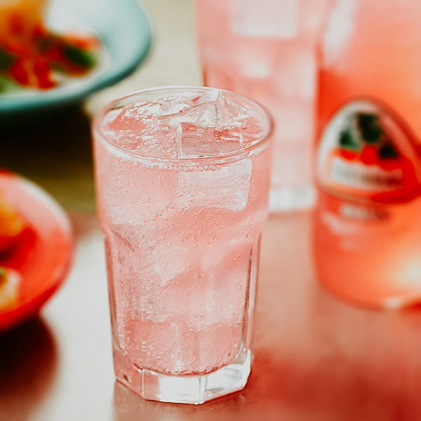 A glass of guava soda with ice, alongside a bottle of the fragrance oil and a plate of food in the background.