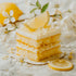 A piece of lemon cake with white frosting garnished with a lemon slice, surrounded by lemon blossoms on a marble surface.