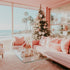 A brightly lit room with a Christmas tree and beach view, featuring a pink sofa, white and pink cushions, and a glass coffee table with a white vase and flowers on it.