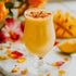 A glass of Mango Lassi, topped with sparkling sugar crystals, with mango slices and flowers in the background.