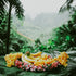 A banana and various colorful flowers arranged on a green leaf, set against a tropical rainforest backdrop.
