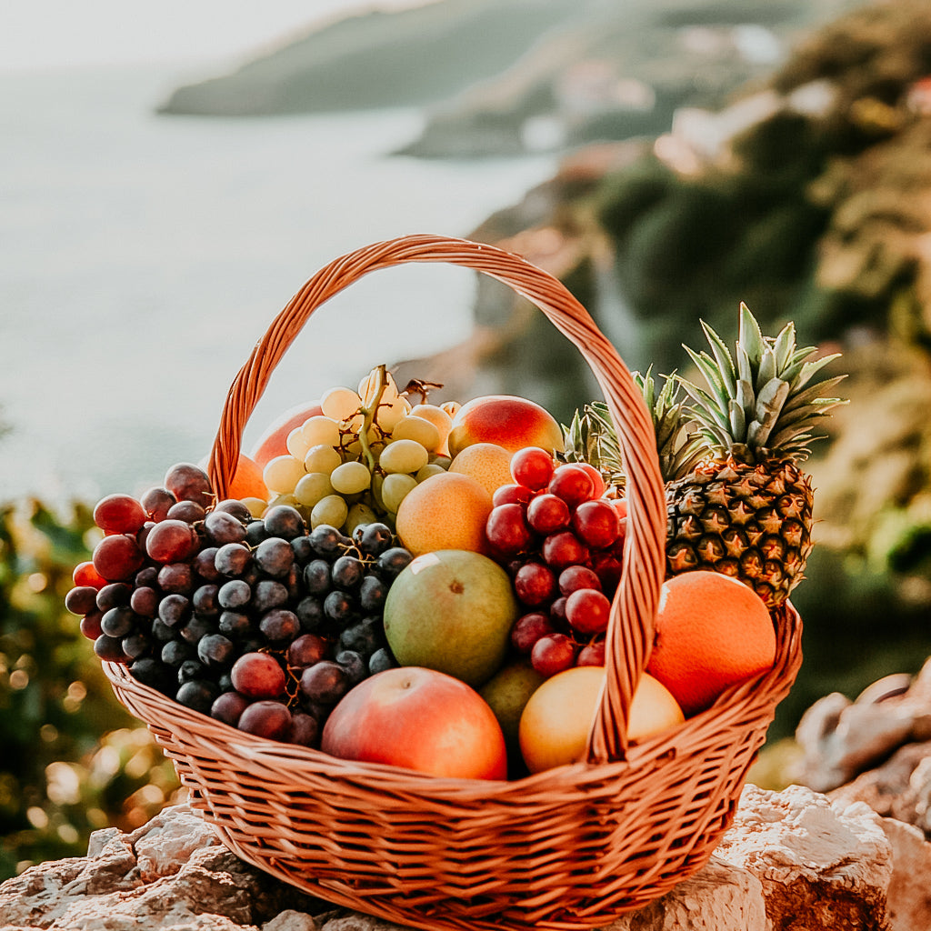 A wicker basket filled with an assortment of fresh fruits including grapes, oranges, apples, and a pineapple, set against a scenic background of a coastal landscape.