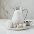 A glass jar filled with milk, on a marble surface, flanked by sugar cubes and a metal cup.