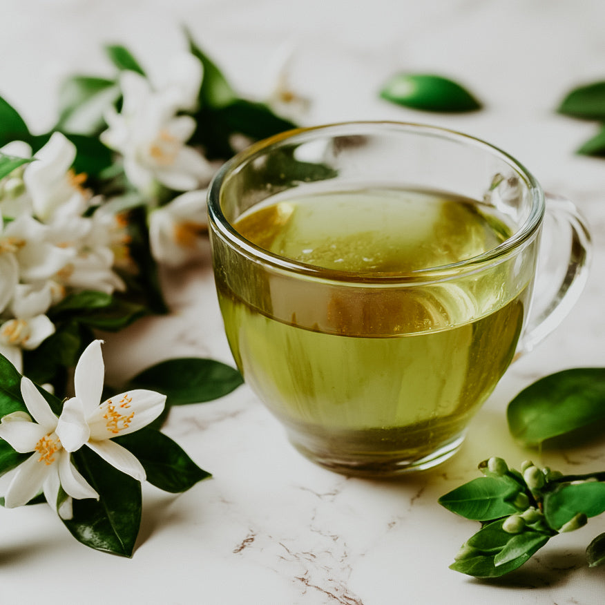 A clear glass cup filled with green tea, surrounded by white flowers and green leaves on a light background.