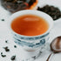 A cup of oolong tea with a tea bag visible in the background, on a white surface with a patterned design.