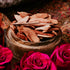 A bowl filled with light brown wooden sticks, possibly representing oud, is placed on a surface with vibrant pink roses around it.