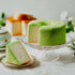 Green sponge cake on a glass cake stand with a slice on a plate, set against a neutral background.
