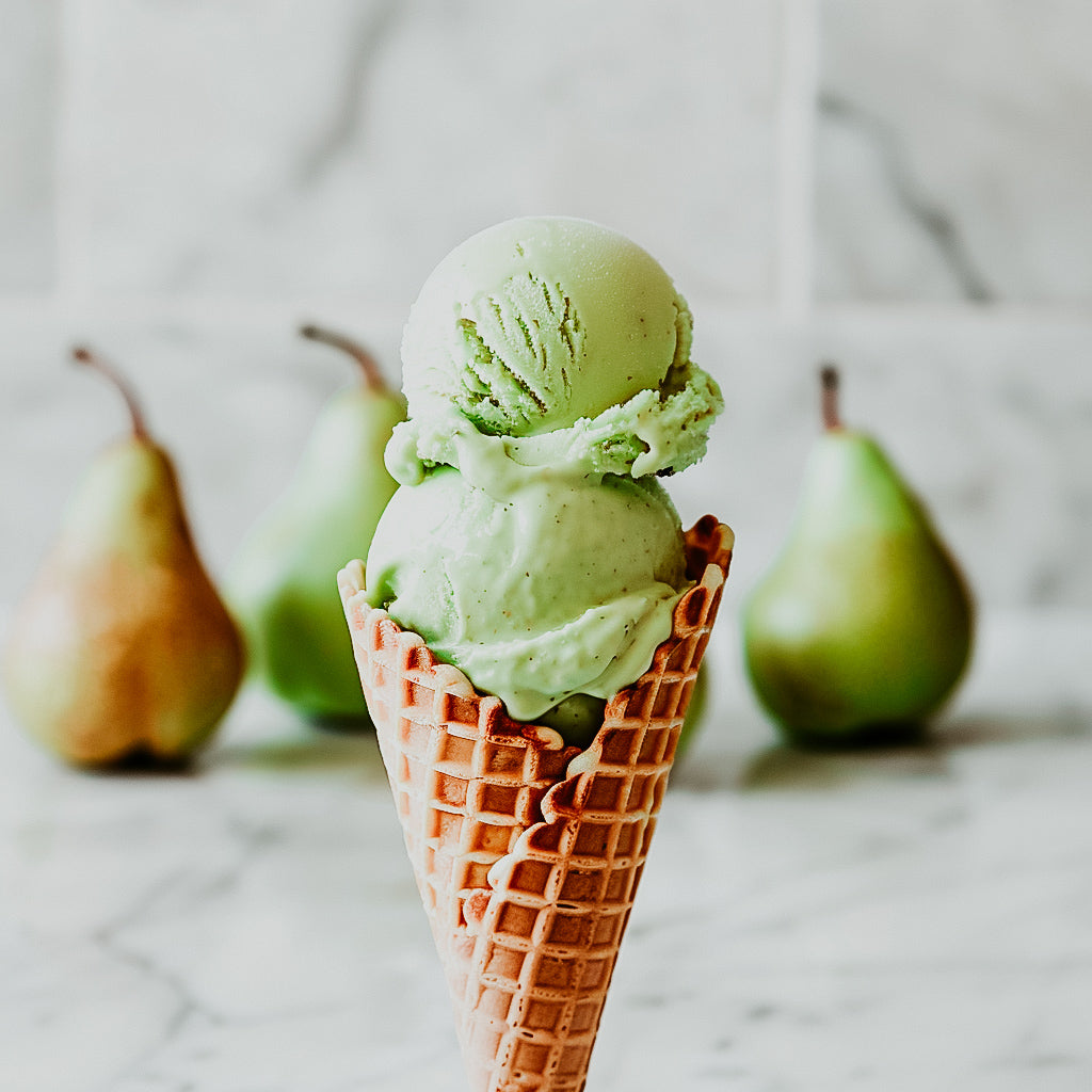 A scoop of green ice cream in a cone, with fresh pears in the background.