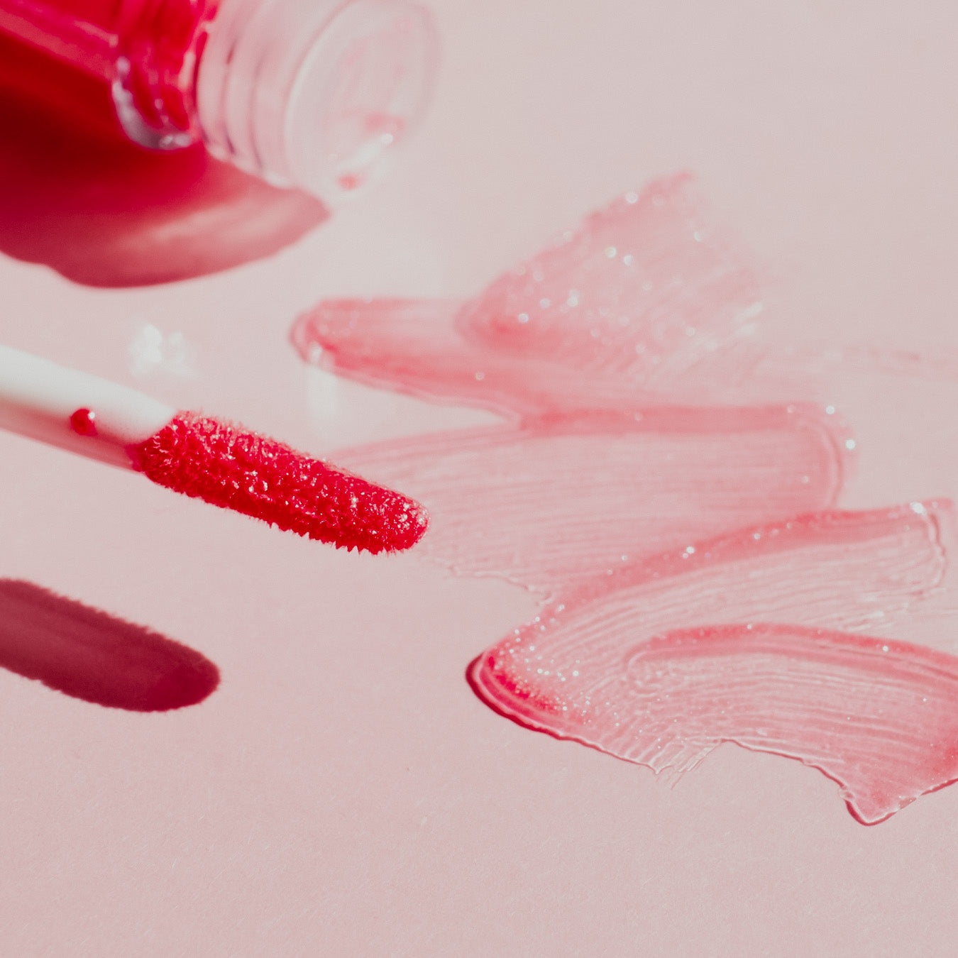 A close-up image of pink lipgloss and its packaging on a pink background, with the lipgloss lid open and a trail of the product leading to the packaging.