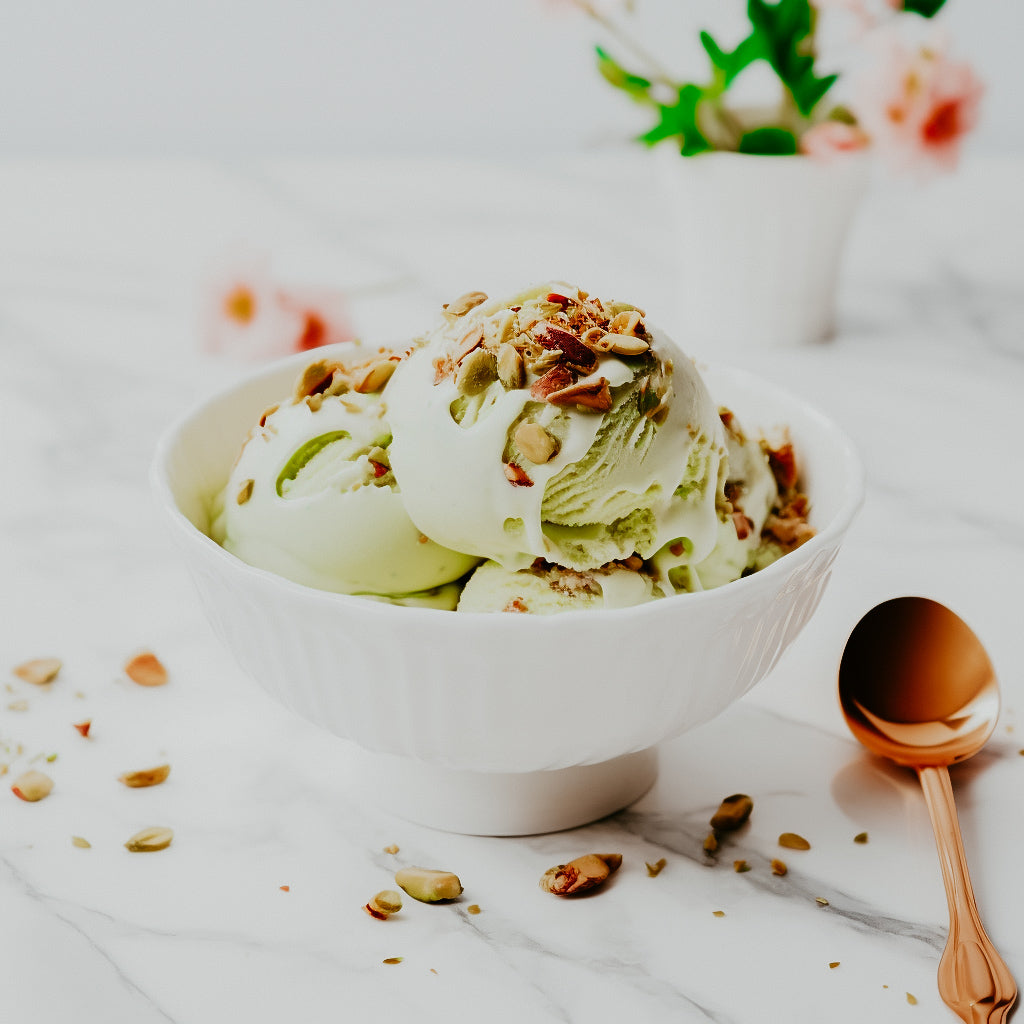 A bowl of pistachio gelato ice cream topped with chopped pistachios, served with a copper spoon on the side, presented on a marble surface with a blurred background.