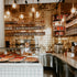 Interior view of a pizzeria with pizzas displayed in the foreground and shelves of ingredients and equipment in the background.