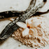 A close-up image of a white powder, resembling vanilla, with a flower at its center, and vanilla pods in the background.