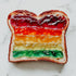 A slice of bread with multicolored layers of jam resembling a rainbow on a white marble surface.