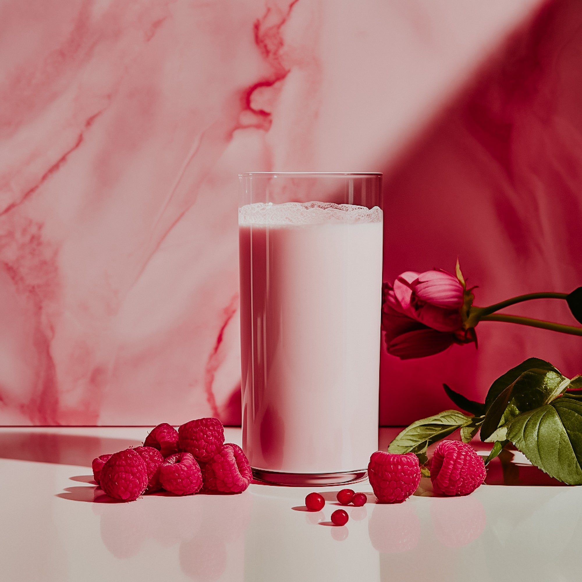 A glass of milk with raspberries and a rose in the foreground, against a pink marble background.