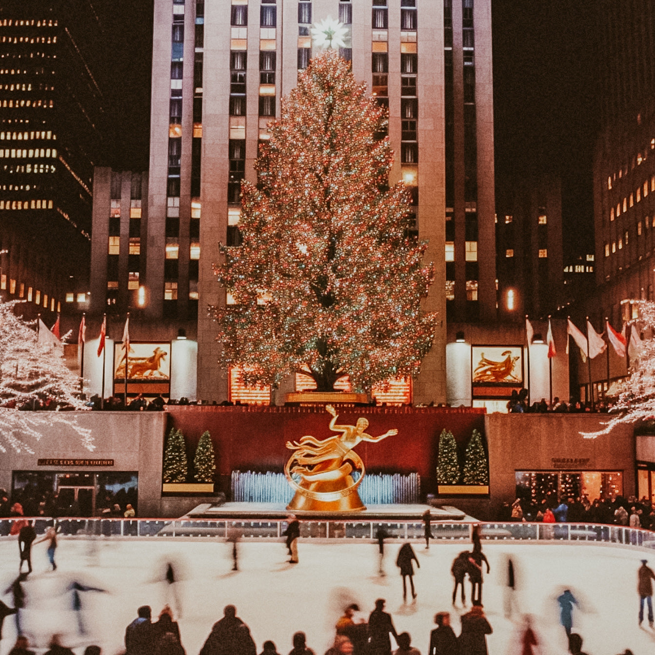 A black and white image of a Rockefeller Center Christmas tree with people skating around it and a red-colored base with a golden statue.