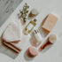 An assortment of self-care items including a fragrance oil bottle, a candle, a loofah, a brush, a bar of soap, a towel, and a small plant on a marble surface.