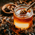 A glass jar filled with honey, with a wooden honey dipper resting on top. The jar is placed on a surface with scattered tobacco leaves around it.