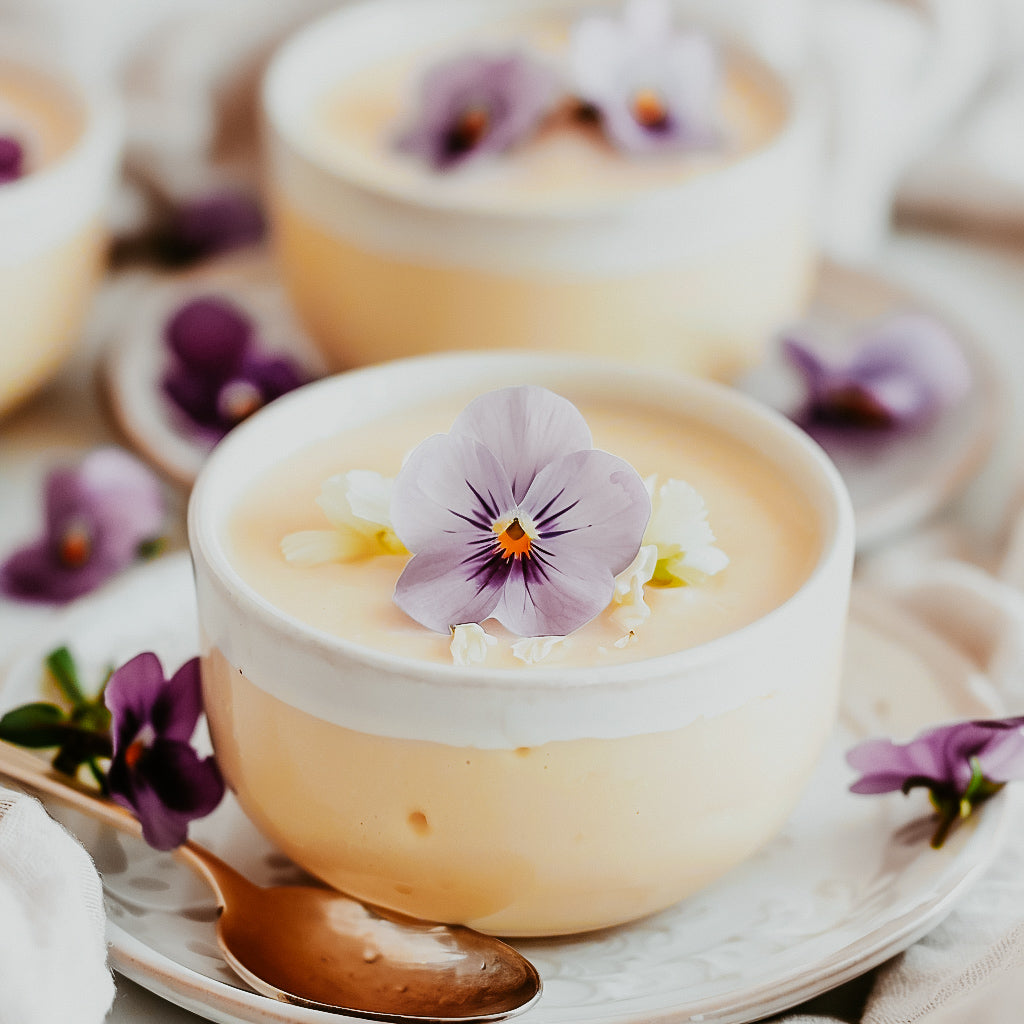 A cup of vanilla custard dessert topped with a flower, served on a white plate with more flowers around it.