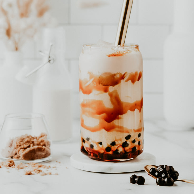 A glass of brown sugar boba milk tea with layers of milk and boba pearls, with a metal straw inserted, displayed on a white surface with a small bowl of brown sugar and blackberries beside it.