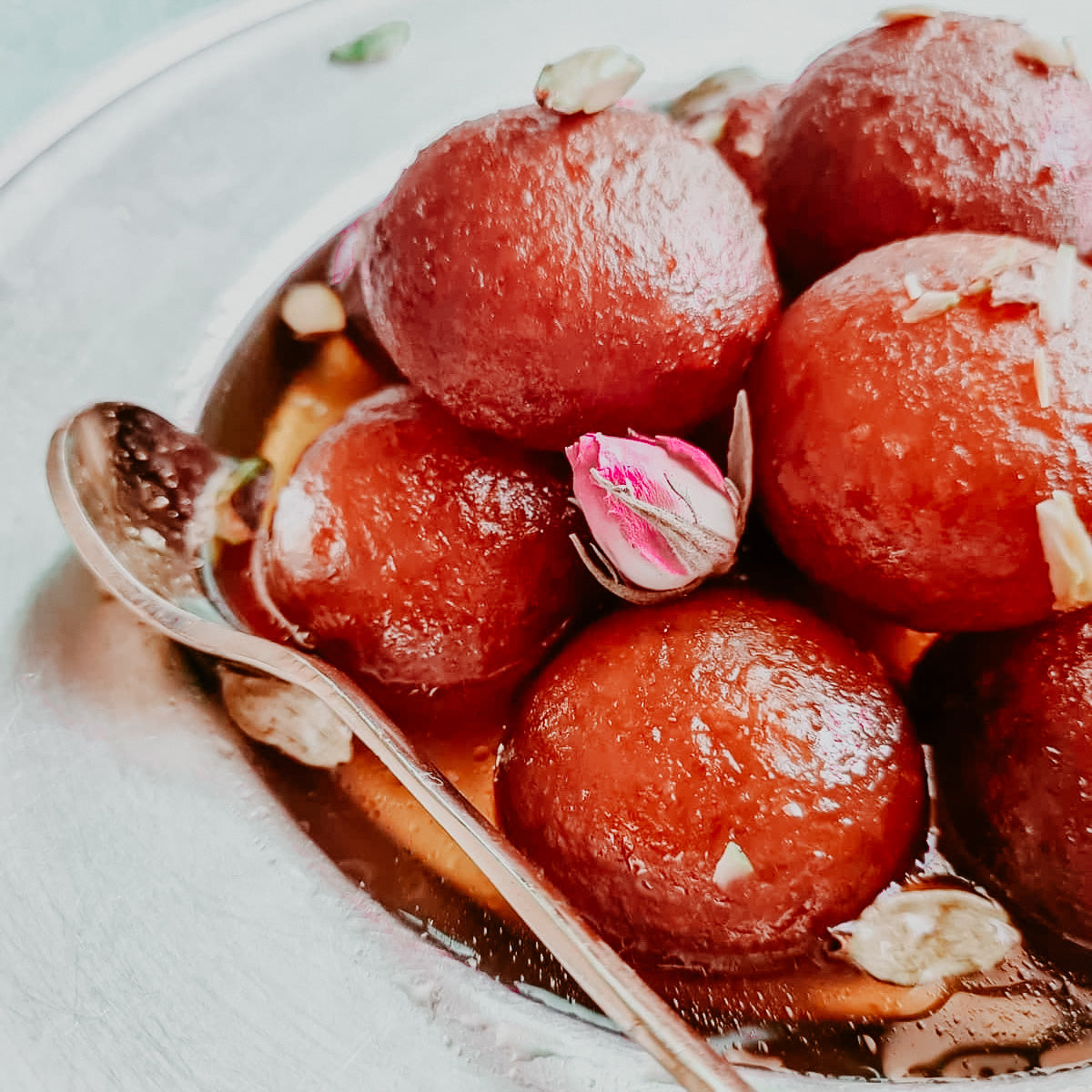 A bowl of Gulab Jamun dessert, consisting of round cake balls immersed in a syrup, garnished with what appears to be rose petals.