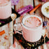 A cup of Kashmiri Chai with milk and spices visible, surrounded by ingredients like rose petals and spices, with a focus on the pink color of the tea.