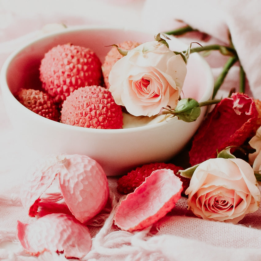 A bowl of pink lychees accompanied by pink roses and petals on a white surface.