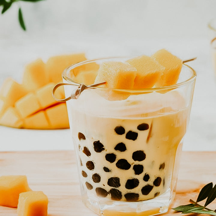 A glass of mango milk tea with boba pearls, alongside slices of mango and a tea bag.