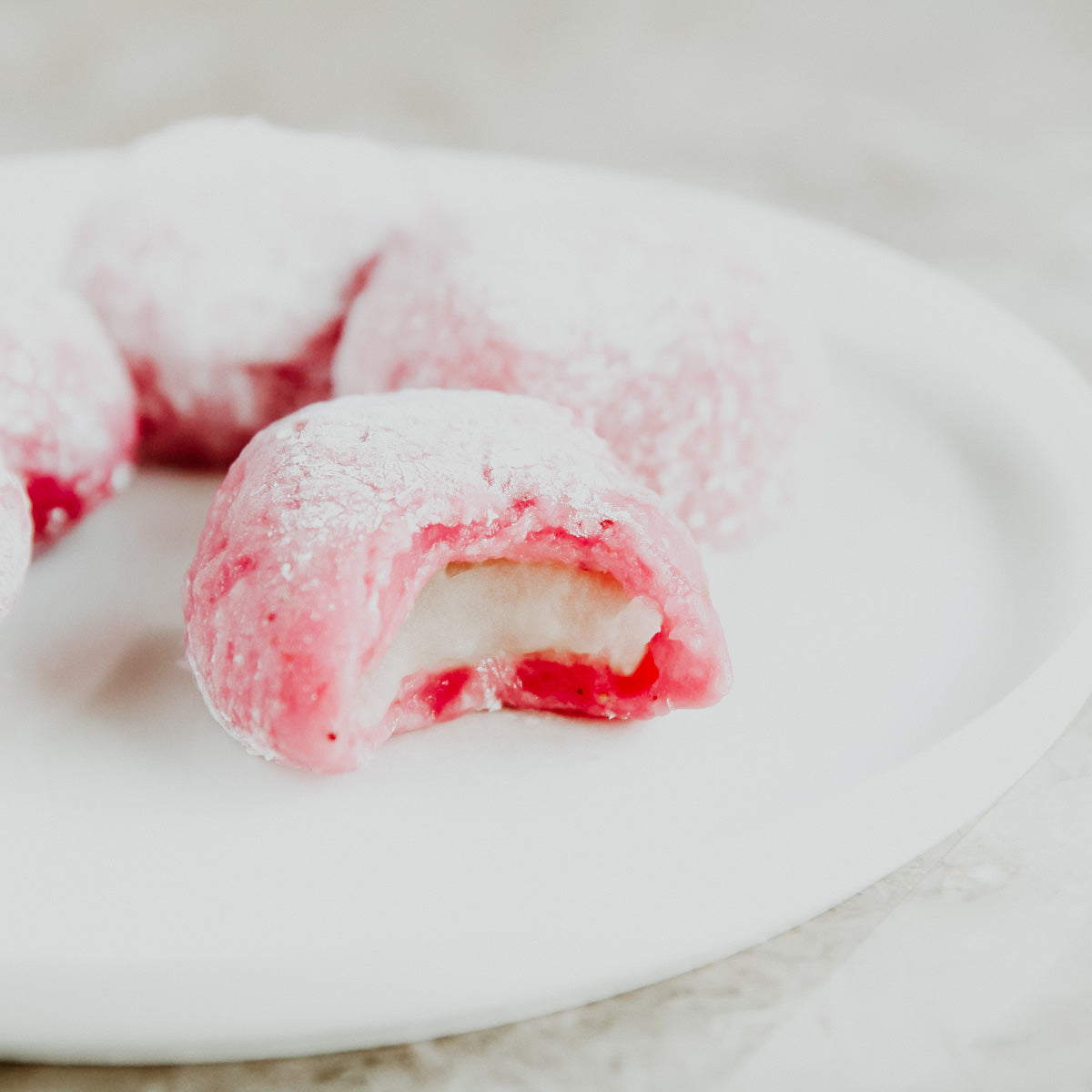 A close-up image of a strawberry mochi with a bite taken out, showing the creamy white filling and the sugary red exterior on a white plate.